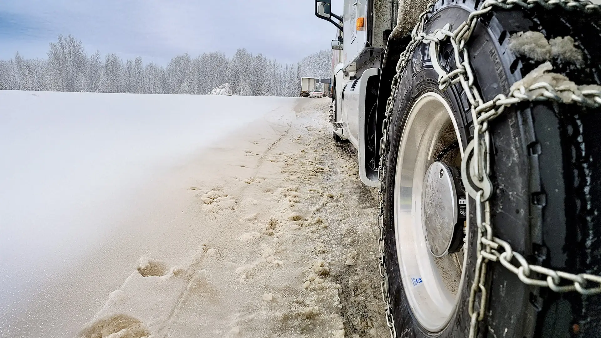 Semi-truck with tire chains driving on a snowy road surrounded by frosted trees, illustrating winter driving safety and tire chain laws for commercial truck drivers.