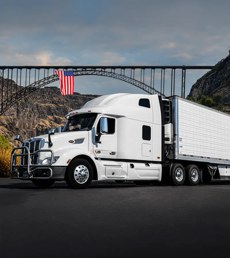 White semi-truck driving across a bridge with an American flag in the background, symbolizing new authority trucking insurance coverage in the United States.